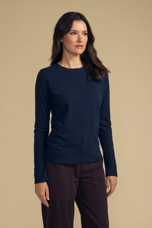 A woman stands against a beige background, wearing the LIZZIE TEE by Velvet by Graham & Spencer—a navy long-sleeve top with a classic crew neckline—paired with dark brown pants, her arms relaxed at her sides.