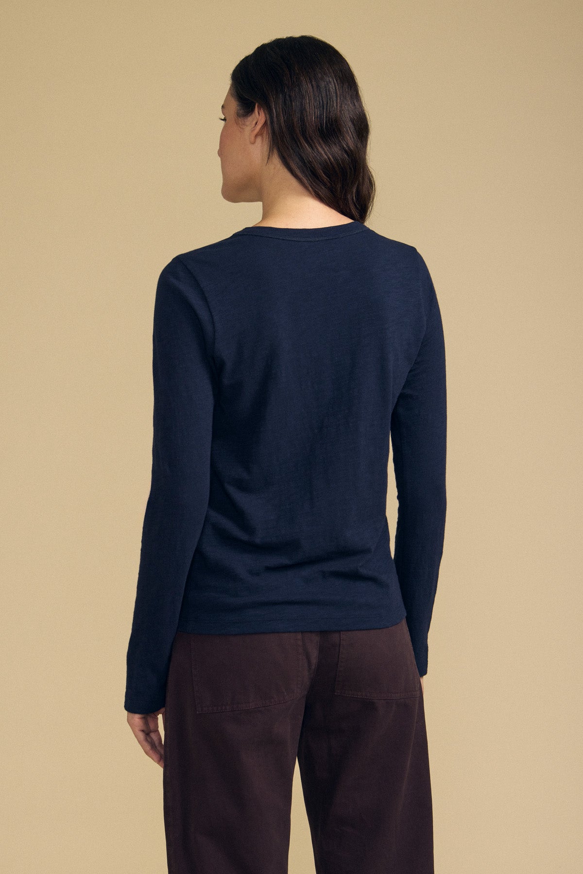 A person with long brown hair, wearing the Velvet by Graham & Spencer LIZZIE TEE—a navy, long sleeve crewneck crafted from textured cotton slub—stands facing away from the camera against a beige background.