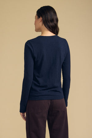A person with long brown hair, wearing the Velvet by Graham & Spencer LIZZIE TEE—a navy, long sleeve crewneck crafted from textured cotton slub—stands facing away from the camera against a beige background.