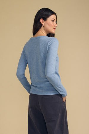 A woman with long brown hair facing sideways wears the Velvet by Graham & Spencer LIZZIE TEE—a light blue, crew neck long sleeve top crafted from textured cotton slub—paired with dark pants, standing against a beige background.