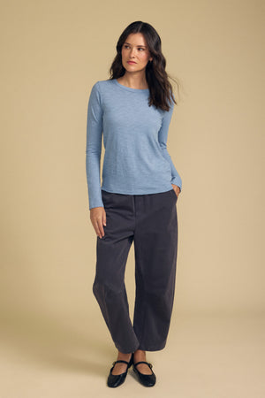 A woman stands against a beige background wearing Velvet by Graham & Spencer’s LIZZIE TEE—a light blue, long sleeve cotton slub top with a crew neckline—paired with loose dark gray pants and black shoes.