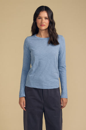 A person stands facing forward against a plain beige background, wearing the Velvet by Graham & Spencer LIZZIE TEE—a light blue, long sleeve cotton slub shirt with a classic crew neckline—and dark pants.