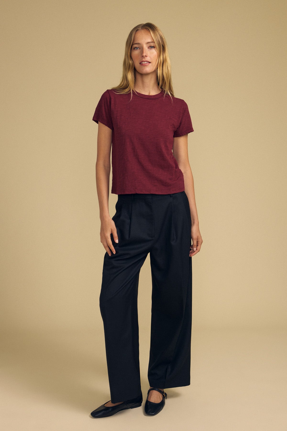 A person stands against a beige background wearing the maroon MARIKA TEE by Velvet by Graham & Spencer, crafted from soft cotton slub fabric. The tee features a relaxed, boxy fit for comfort and effortless style, and is paired with black wide-leg trousers and black shoes.-41929412608193