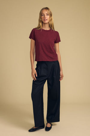A person stands against a beige background wearing the maroon MARIKA TEE by Velvet by Graham & Spencer, crafted from soft cotton slub fabric. The tee features a relaxed, boxy fit for comfort and effortless style, and is paired with black wide-leg trousers and black shoes.