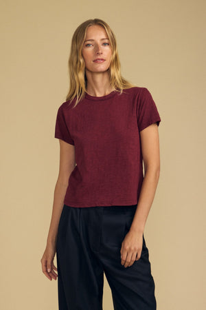 A woman wearing a short-sleeve burgundy MARIKA TEE by Velvet by Graham & Spencer, crafted from soft cotton slub in a relaxed boxy fit, stands against a plain beige background.