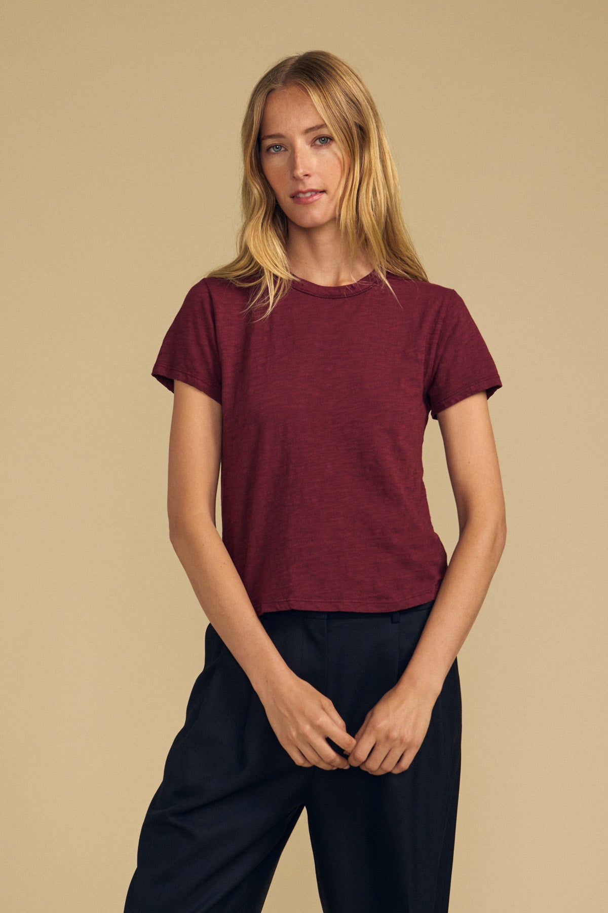 A woman with long blonde hair stands against a plain beige background, wearing the Velvet by Graham & Spencer MARIKA TEE in maroon—a short-sleeve top featuring a relaxed, boxy fit—paired with black pants.