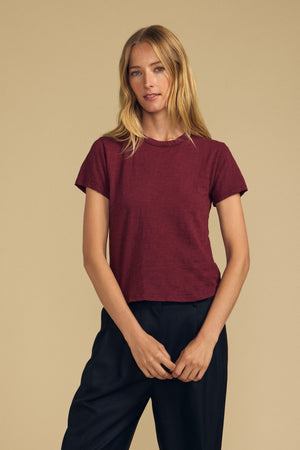A woman with long blonde hair stands against a plain beige background, wearing the Velvet by Graham & Spencer MARIKA TEE in maroon—a short-sleeve top featuring a relaxed, boxy fit—paired with black pants.
