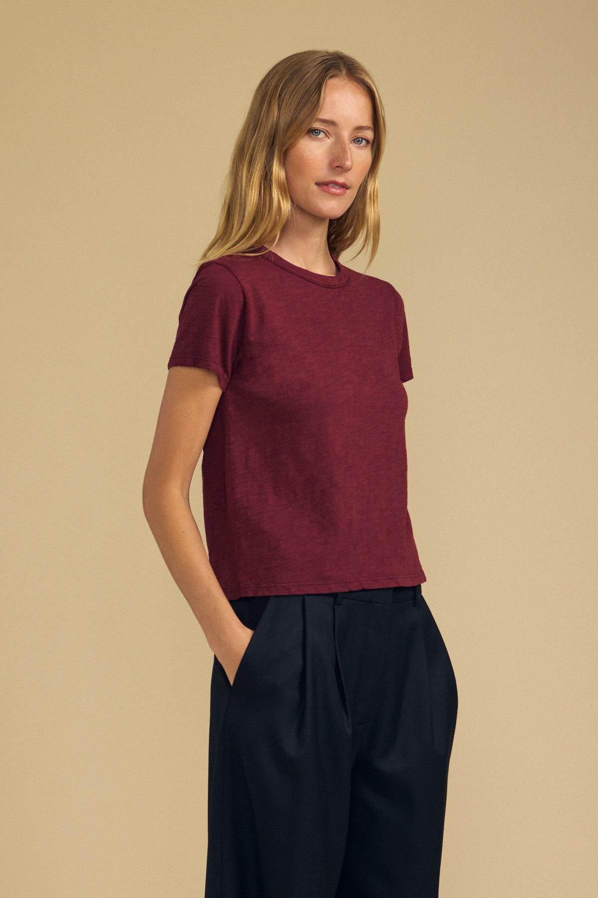 A person with long blonde hair wears a short-sleeve maroon MARIKA TEE by Velvet by Graham & Spencer in a relaxed boxy fit, crafted from soft cotton slub, paired with black pants, standing against a plain beige background with hands in pockets.