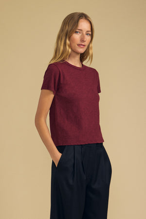 A person with long blonde hair wears a short-sleeve maroon MARIKA TEE by Velvet by Graham & Spencer in a relaxed boxy fit, crafted from soft cotton slub, paired with black pants, standing against a plain beige background with hands in pockets.