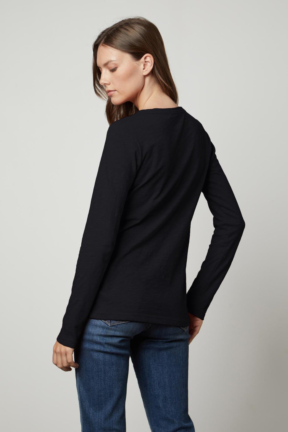   The back view of a woman wearing jeans and a black LIZZIE ORIGINAL SLUB LONG SLEEVE TEE made by Velvet by Graham & Spencer, made of cotton. 