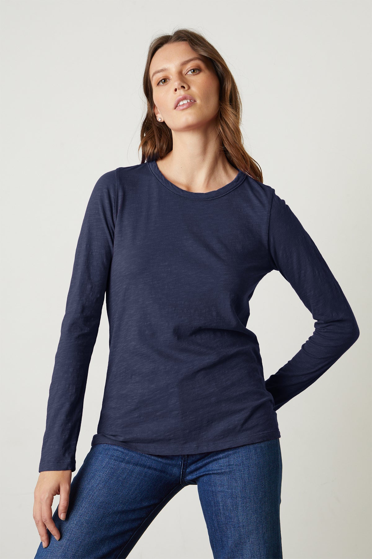 A woman models the LIZZIE TEE by Velvet by Graham & Spencer— a navy, long-sleeve crew neck—paired with blue jeans, standing with one hand on her hip against a plain light background.-39434505224385