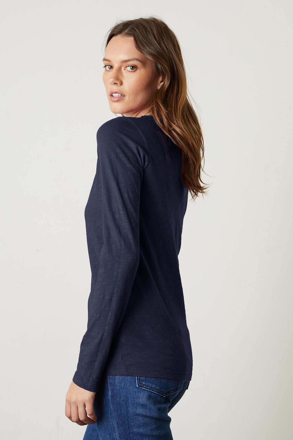 A woman with long brown hair stands sideways against a plain light background, wearing the Velvet by Graham & Spencer LIZZIE TEE in navy textured cotton slub and blue jeans, looking over her shoulder.-39434505257153