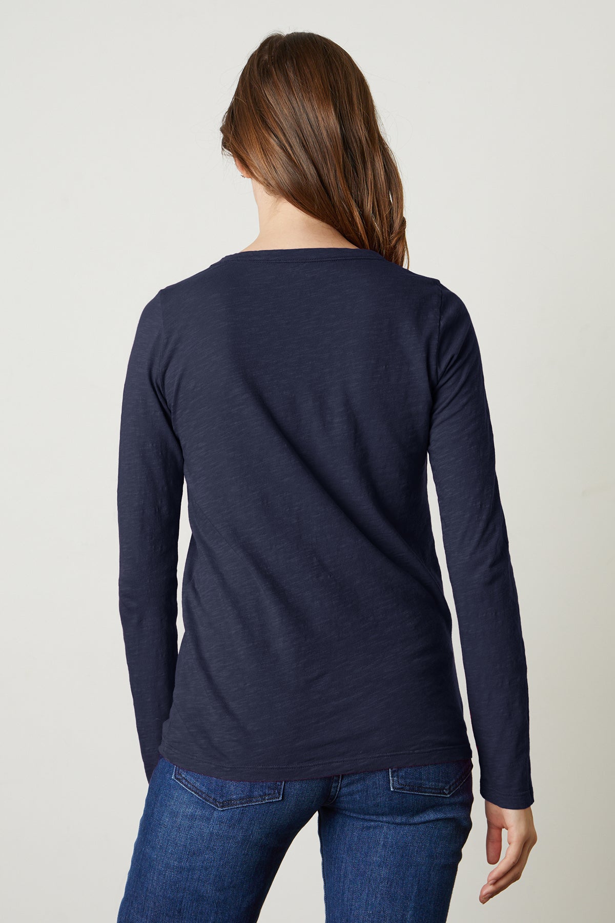 A woman with long brown hair, wearing the Velvet by Graham & Spencer LIZZIE TEE in navy and blue jeans, stands facing away from the camera against a plain background.-39434505289921