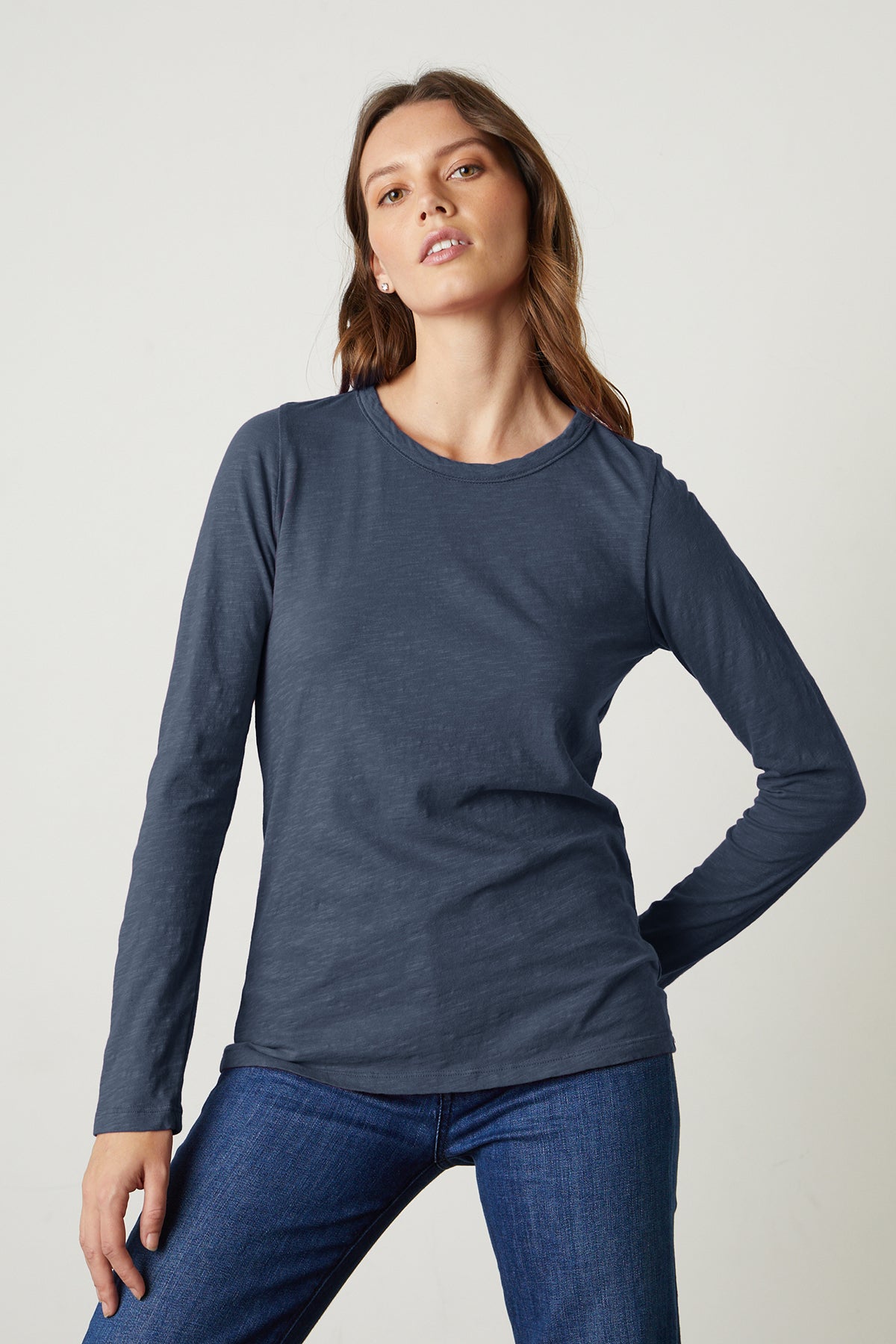   A woman with long hair is standing against a pale background, wearing the Velvet by Graham & Spencer LIZZIE TEE in navy-blue, which features long sleeves and a classic crew neckline, along with dark blue jeans. 