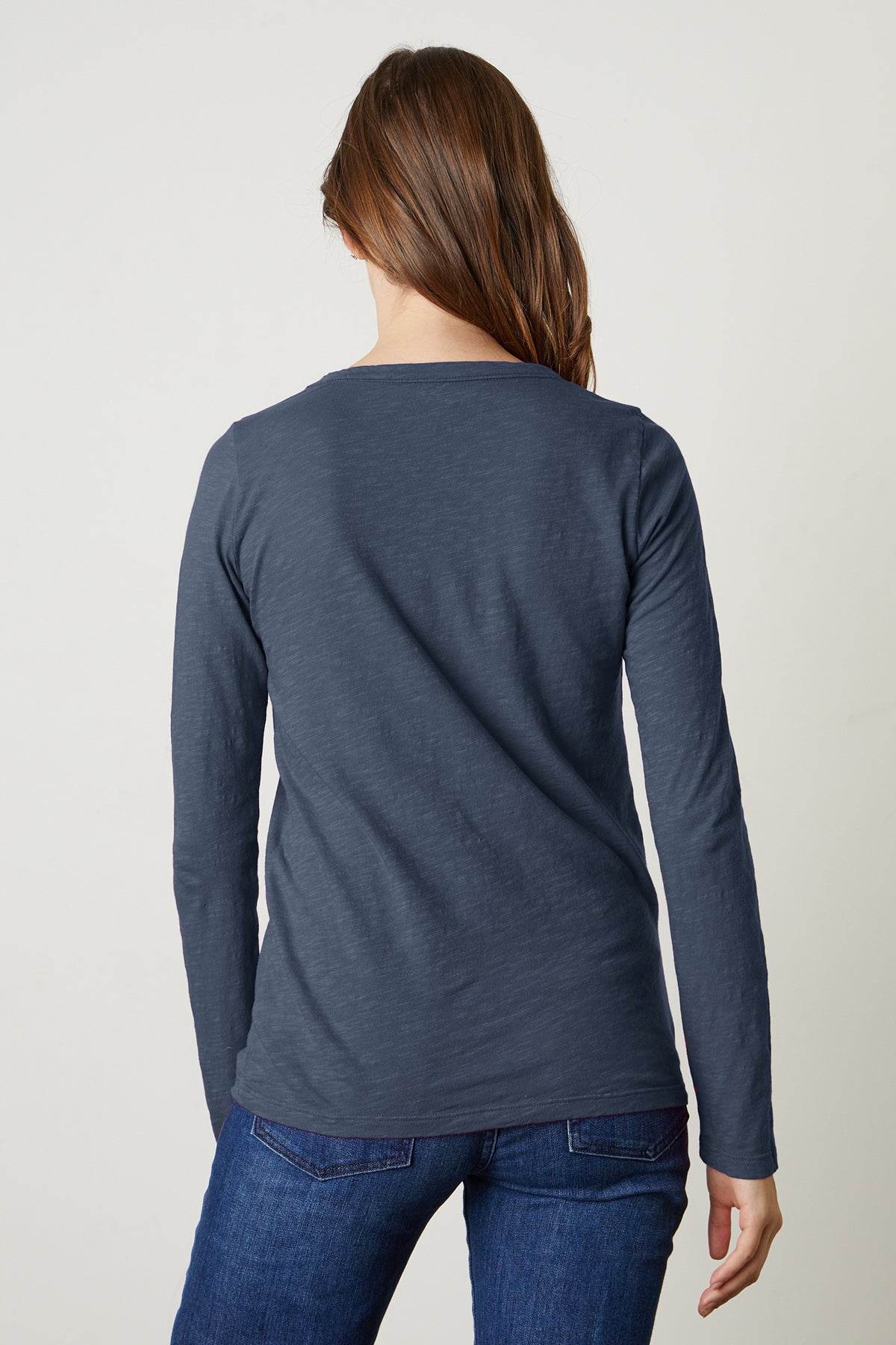   A person with long brown hair is shown from the back, wearing the dark blue LIZZIE TEE by Velvet by Graham & Spencer, featuring long sleeves and a classic crew neckline, paired with blue jeans, standing against a plain background. 