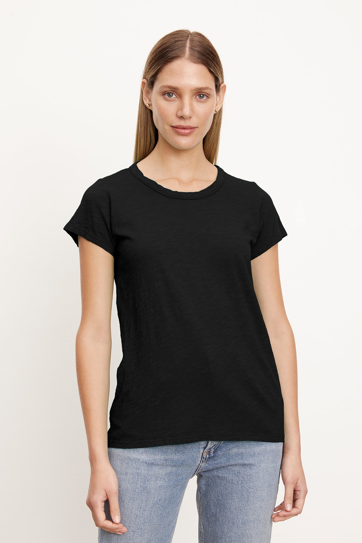   A person with long light brown hair is wearing a plain black TILLY TEE by Velvet by Graham & Spencer made of soft cotton slub and light blue jeans, standing against a white background. 