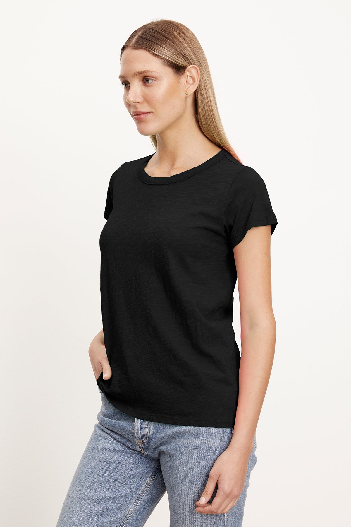 A person with long hair stands with a slight smile, wearing a black TILLY TEE by Velvet by Graham & Spencer made of soft cotton slub and light blue jeans against a plain white background.-37355955847361