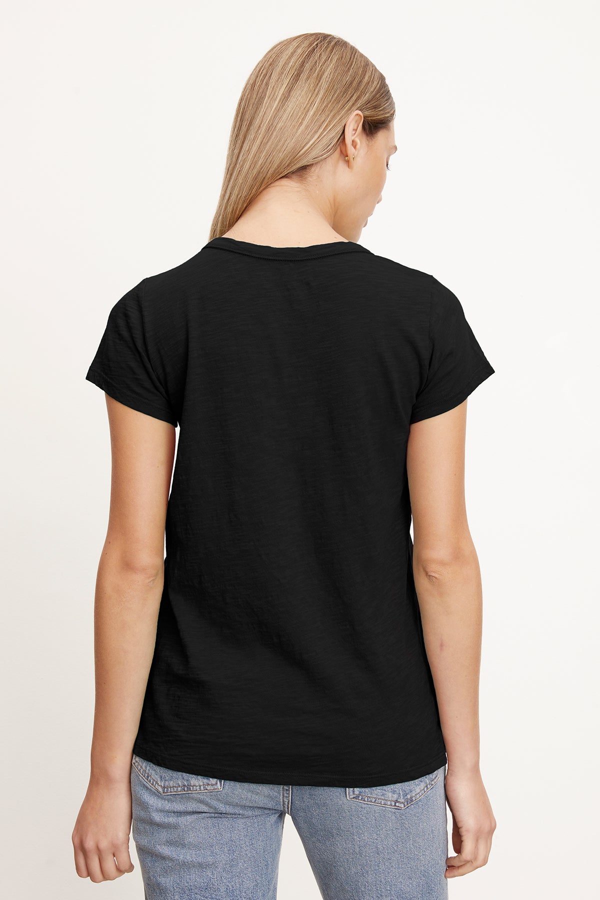   A person with long blonde hair wearing a black TILLY TEE by Velvet by Graham & Spencer and blue jeans is facing away, showcasing the California classic back view. 