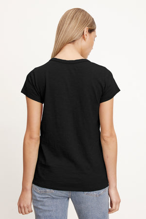 A person with long blonde hair wearing a black TILLY TEE by Velvet by Graham & Spencer and blue jeans is facing away, showcasing the California classic back view.