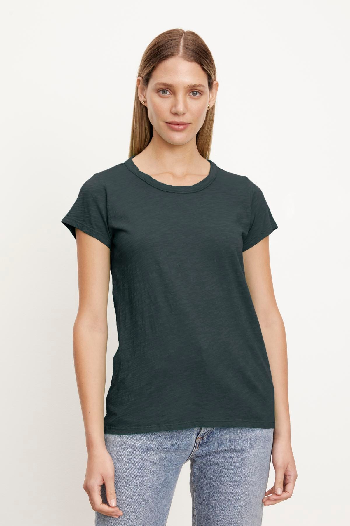   Woman with straight, shoulder-length hair wearing a dark green TILLY TEE by Velvet by Graham & Spencer made from soft cotton slub and light blue jeans, standing against a plain white background. 