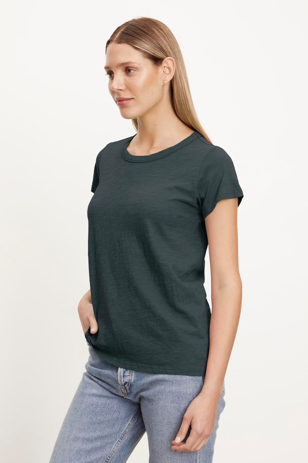   A woman with long blonde hair is wearing a dark green Velvet by Graham & Spencer TILLY TEE and light blue jeans, standing with one hand in her pocket against a plain white background. 