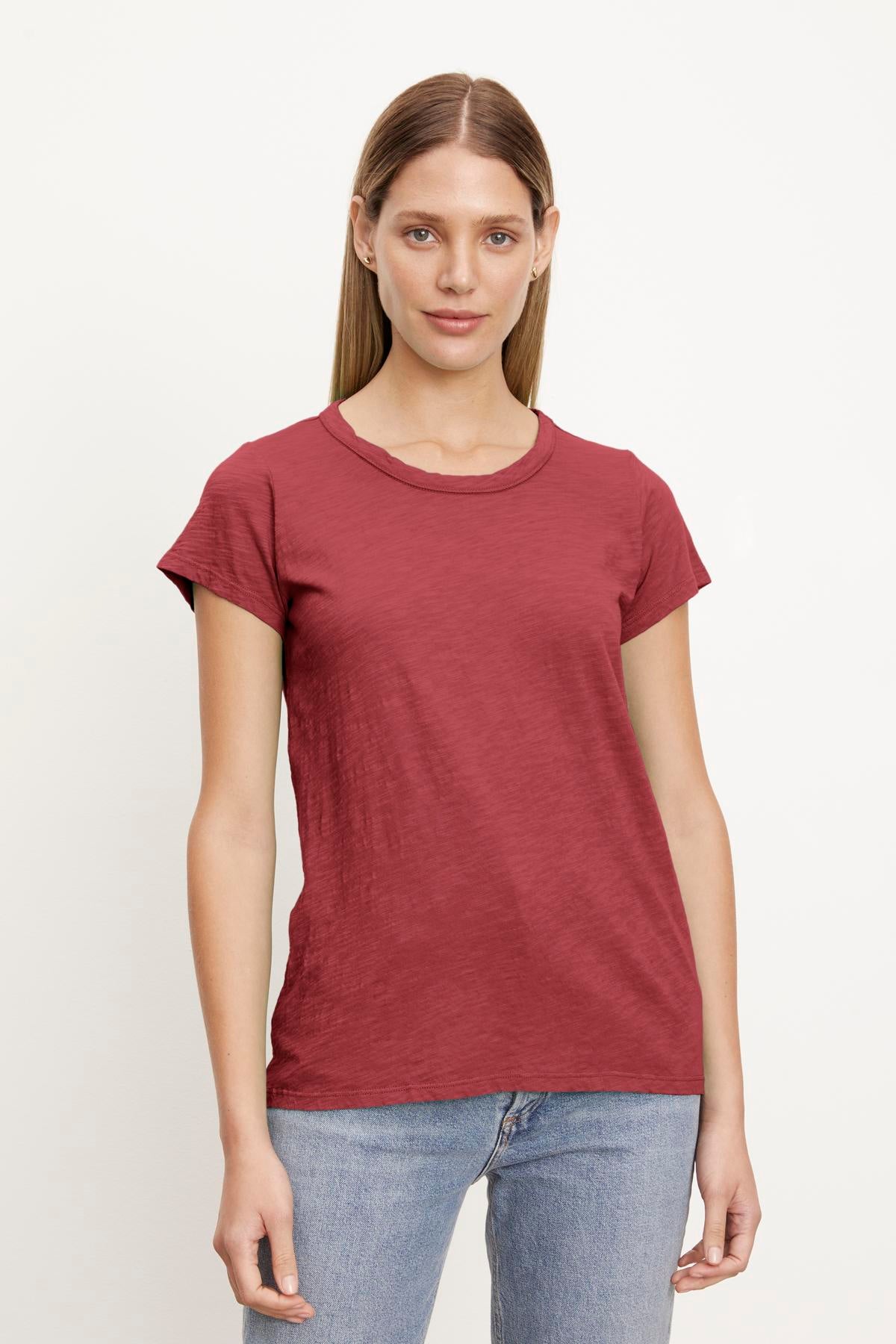   A woman stands against a plain background, wearing a short-sleeved, red TILLY TEE made of soft cotton slub from Velvet by Graham & Spencer and blue jeans, with a neutral expression and straight, long hair. 