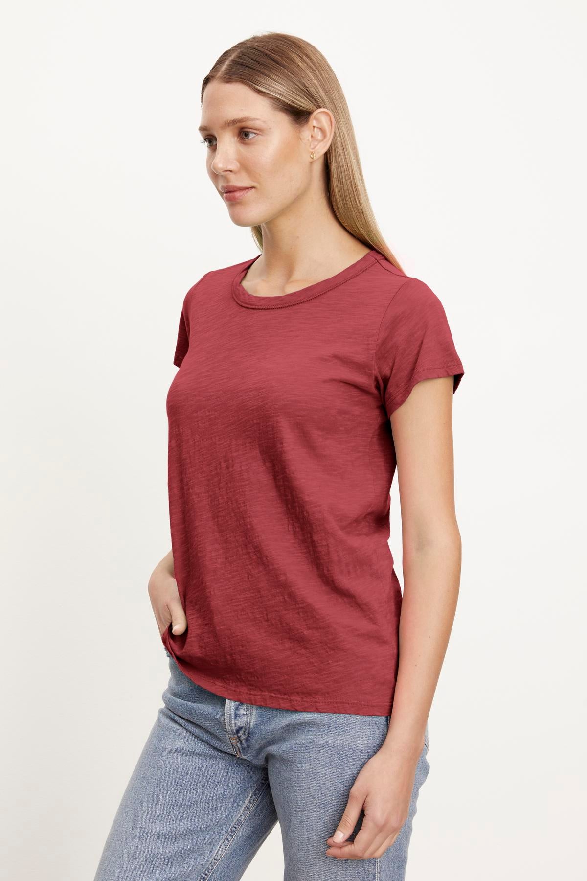   A person with long hair wearing a TILLY TEE by Velvet by Graham & Spencer, a soft cotton slub, red short-sleeved crew neck tee and light blue jeans stands with one hand in their pocket against a plain white background. 