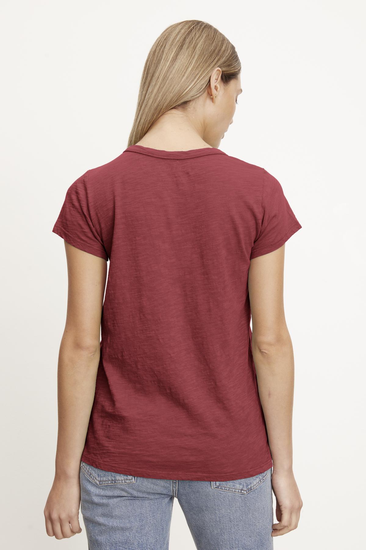   A person with long blonde hair, wearing a red TILLY TEE by Velvet by Graham & Spencer and blue jeans, is standing with their back to the camera against a plain white background. 