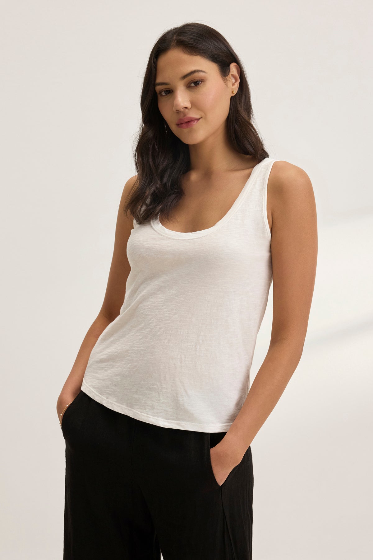 A woman in a white AYA TANK TOP by Velvet by Graham & Spencer and black pants stands with hands in pockets against a plain background. Crafted from textured cotton slub, her sleeveless top with a scoop neckline is perfect for effortless layering.-38957693272257