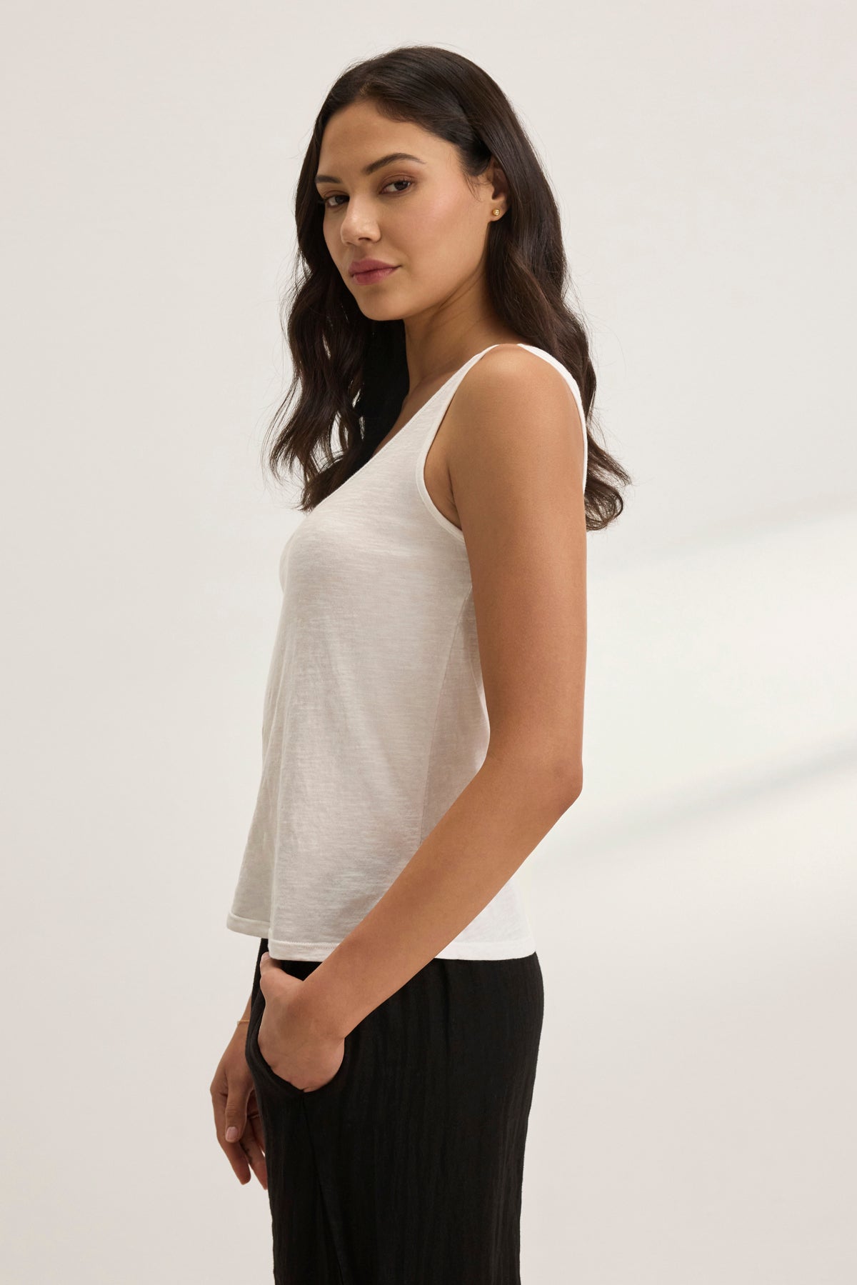 A person with long dark hair stands sideways against a plain background, wearing the AYA TANK TOP from Velvet by Graham & Spencer and black pants, with one hand in their pocket.-38957693239489