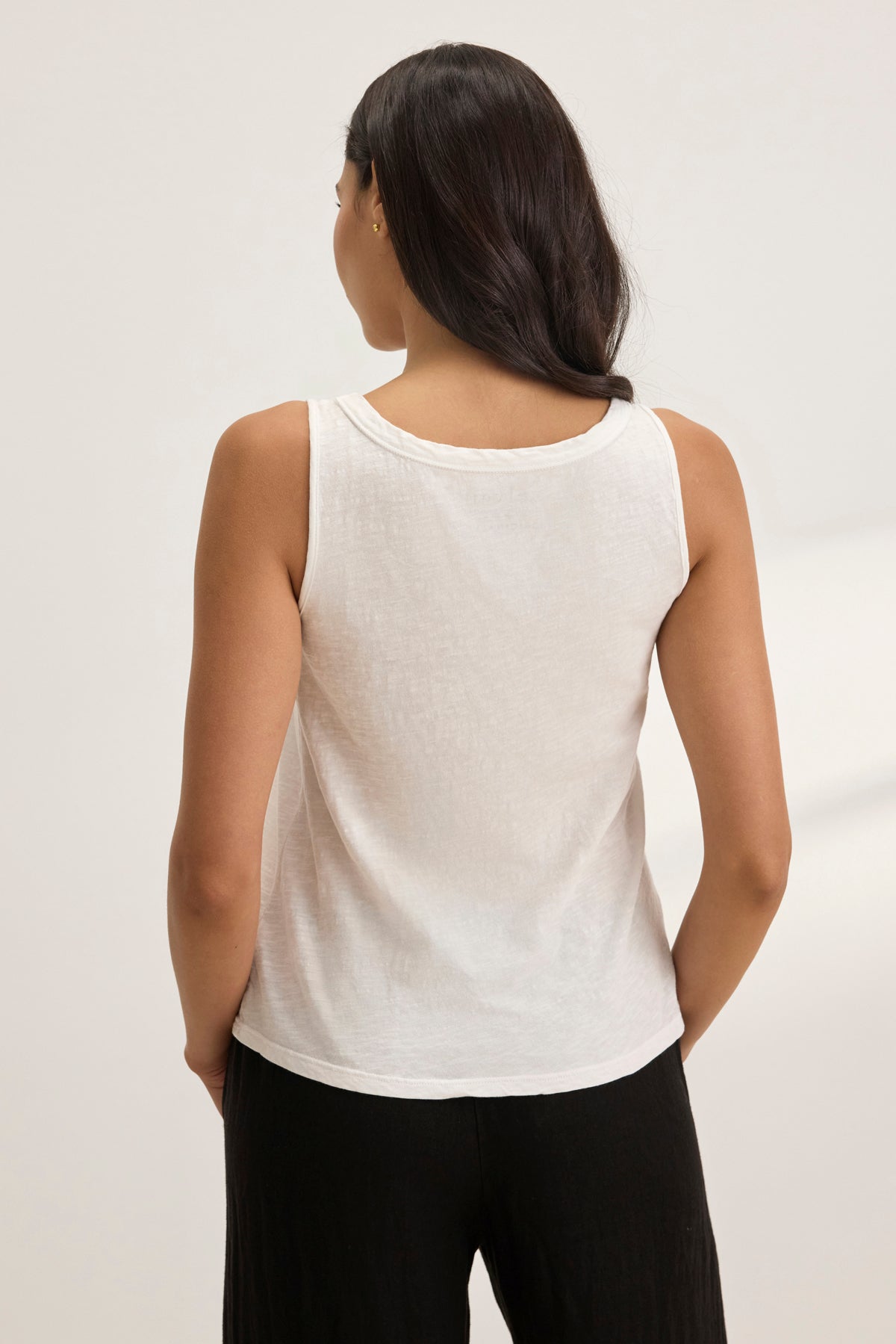 A woman with long dark hair is facing away, highlighting the AYA TANK TOP by Velvet by Graham & Spencer. The white sleeveless top features a scoop neckline and textured cotton slub, paired with black pants against a plain background for an effortless minimalist look.-38957693206721