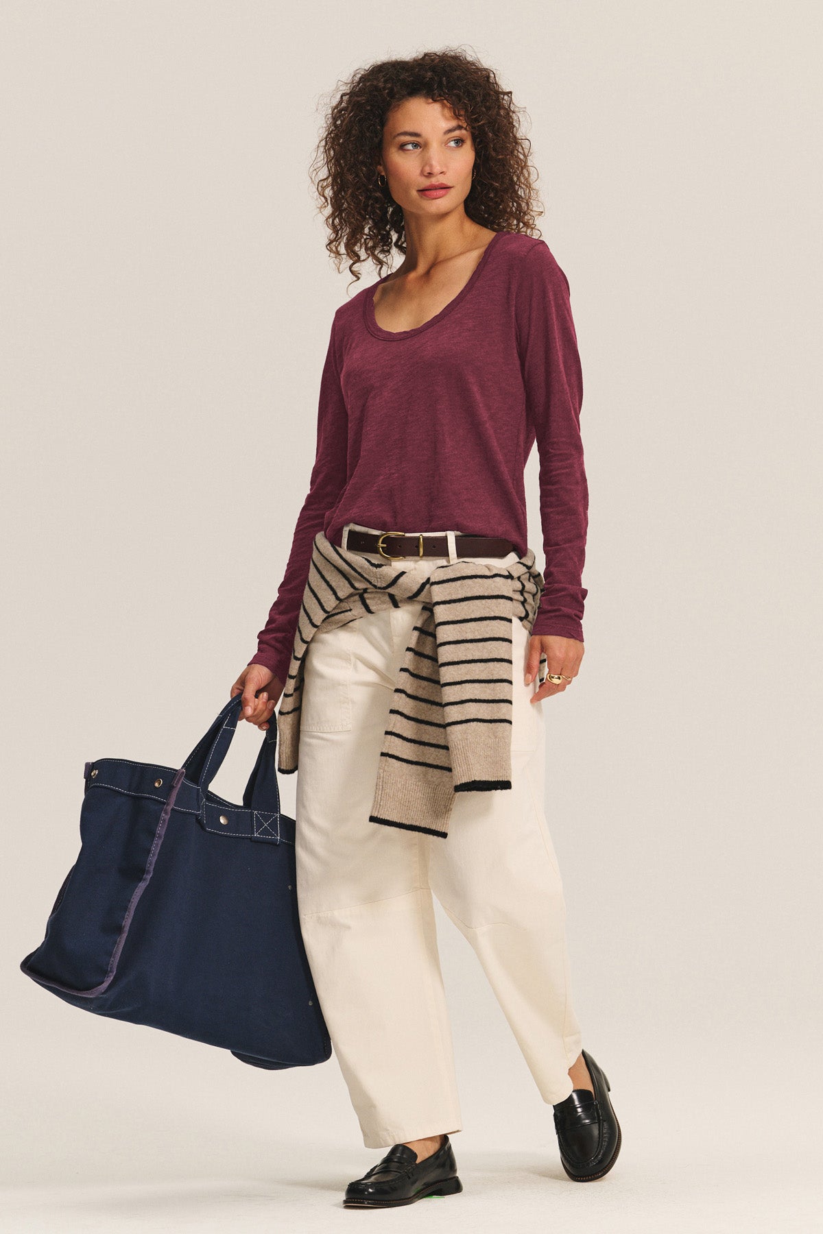 A woman in the BECKY TEE by Velvet by Graham & Spencer (maroon, relaxed scoop neck, long sleeves), beige pants, black shoes, and a striped sweater at her waist holds a large navy tote.