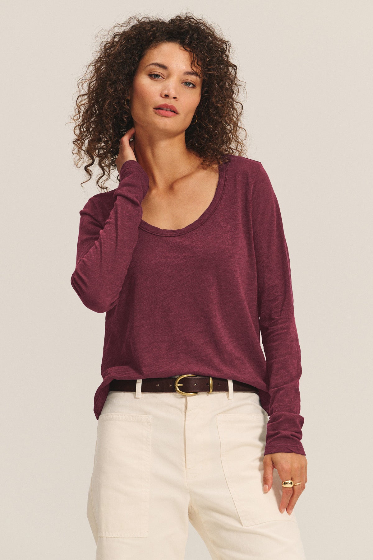 A woman wears the BECKY TEE by Velvet by Graham & Spencer in maroon with a relaxed scoop neck, paired with high-waisted white pants and a dark belt, standing against a plain light background.