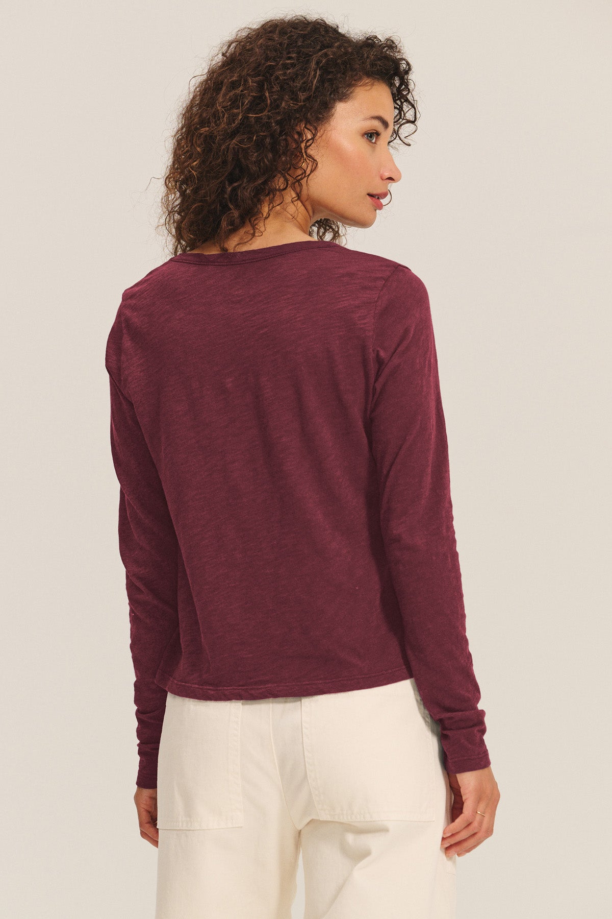 A person is seen from behind wearing the Velvet by Graham & Spencer BECKY TEE, a maroon long-sleeve scoop neck top, paired with white pants against a plain light background.