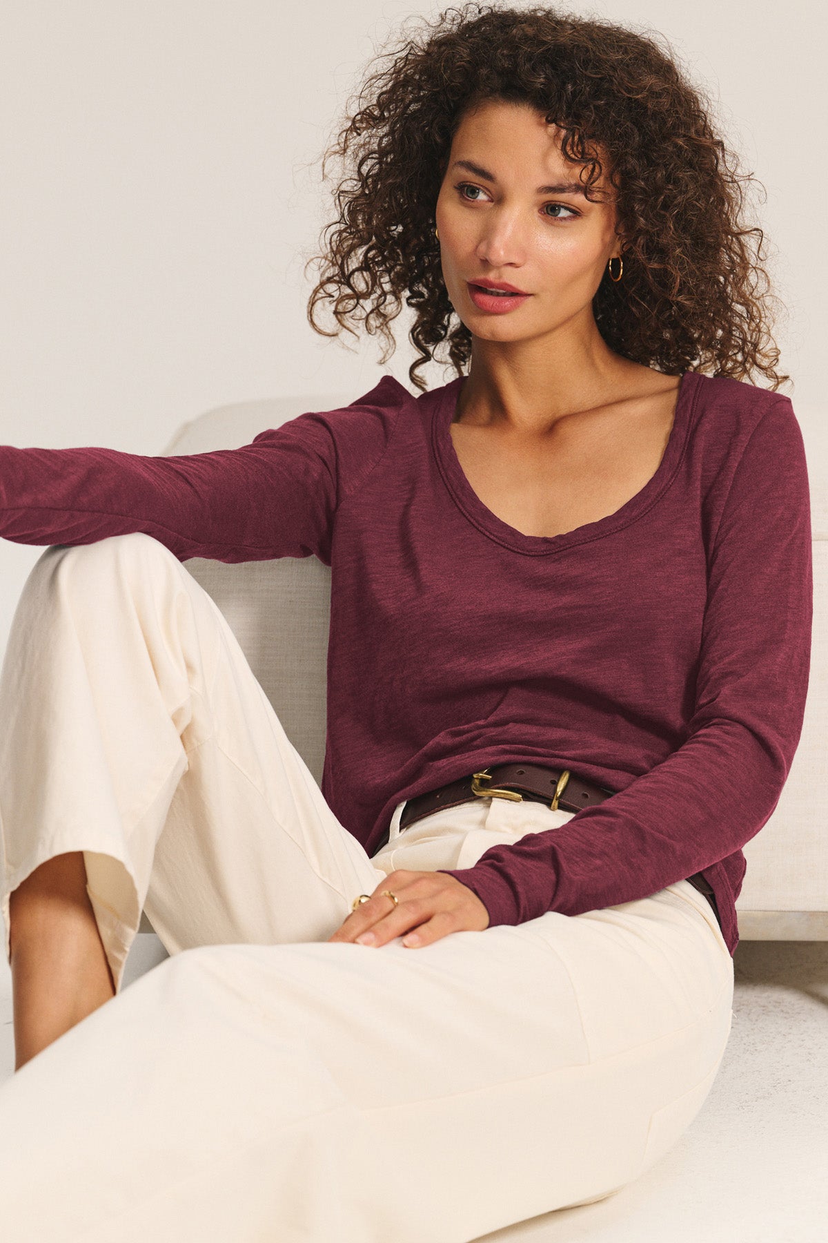 A woman with curly hair sits on a light-colored couch, looking to the side, wearing the Velvet by Graham & Spencer BECKY TEE in maroon and cream-colored pants.