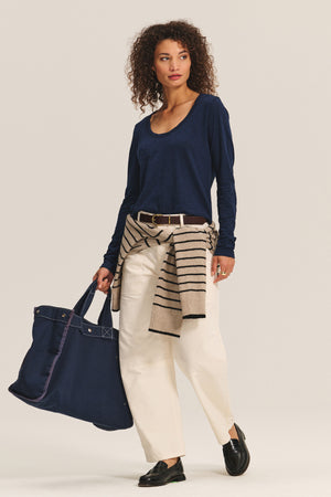 A woman wears the BECKY TEE by Velvet by Graham & Spencer, a navy relaxed scoop neck long-sleeve top, paired with cream pants, black shoes, a striped sweater tied at her waist, and carries a large navy tote bag.