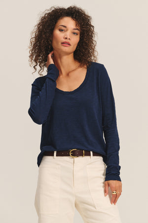 A woman wears the BECKY TEE by Velvet by Graham & Spencer—a navy cotton slub long-sleeve tee with a relaxed scoop neck—tucked into high-waisted cream pants and finished with a brown belt, standing against a plain light background.
