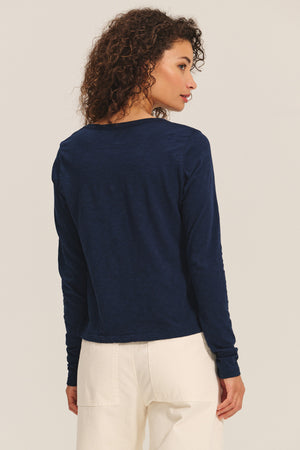 A woman with curly hair stands with her back to the camera, wearing the BECKY TEE by Velvet by Graham & Spencer in navy blue and off-white pants against a plain background.