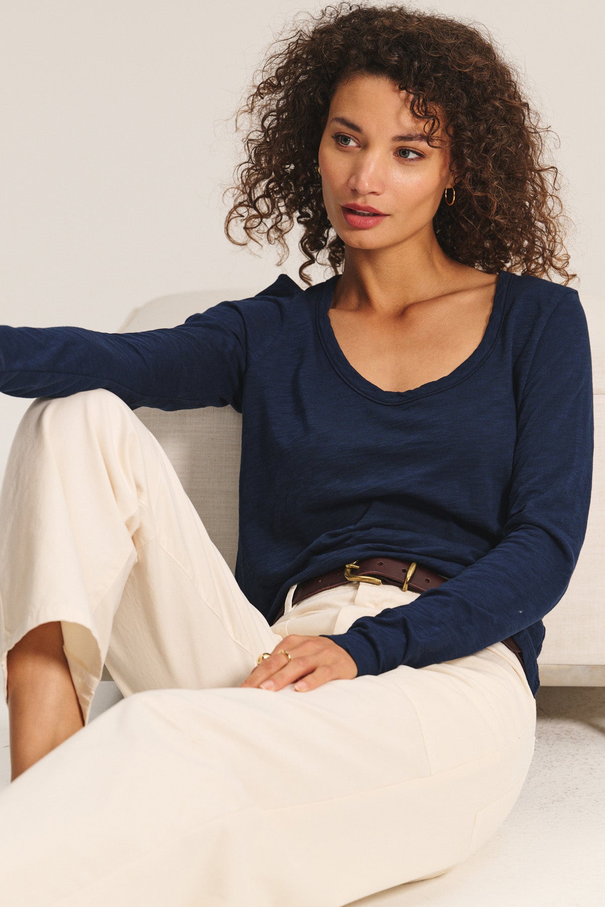 A woman with curly hair sits on a light-colored sofa, wearing the BECKY TEE by Velvet by Graham & Spencer—a navy cotton slub long-sleeve top with a relaxed scoop neck—paired with cream pants and a brown belt.