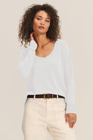 A woman with curly hair wears the BECKY TEE by Velvet by Graham & Spencer, a white cotton slub long-sleeve top tucked into high-waisted cream pants with a brown belt, standing against a neutral background.