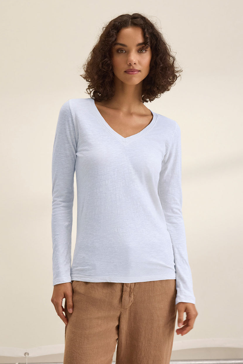 A woman with curly brown hair wears the BLAIRE TEE by Velvet by Graham & Spencer—a light blue, long-sleeve V-neck shirt in textured cotton slub—paired with tan pants against a plain background, ideal for everyday wear.