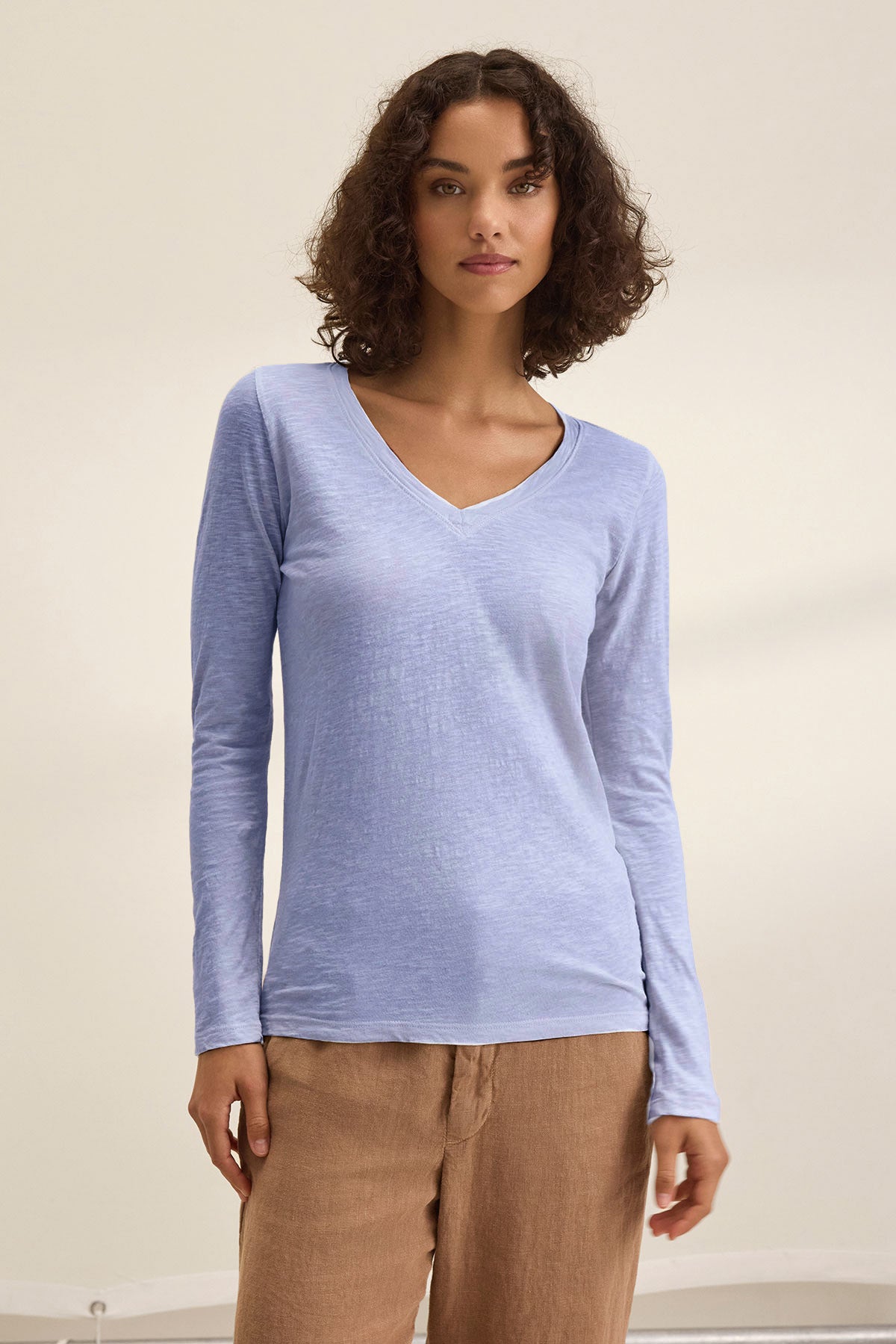 A woman with curly hair wears the Velvet by Graham & Spencer BLAIRE TEE, a light blue, long-sleeve cotton slub v-neck, paired with tan pants against a plain light background.-41506761769153