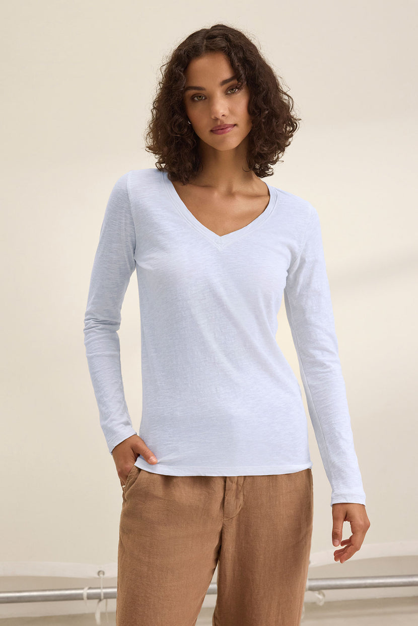 A person with short curly hair wears the BLAIRE TEE by Velvet by Graham & Spencer—a light blue, long-sleeve V-neck shirt in textured cotton slub—paired with tan pants, standing against a plain background with one hand in a pocket.