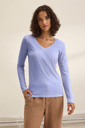 A woman faces forward against a plain background, wearing the BLAIRE TEE by Velvet by Graham & Spencer—a light blue cotton slub, long sleeve v-neck tee—paired with beige pants.