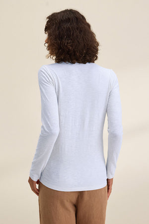 A person with curly hair wears the Velvet by Graham & Spencer BLAIRE TEE, a long-sleeved white v-neck shirt, paired with brown pants and photographed from the back against a neutral backdrop—perfect for effortless everyday style.