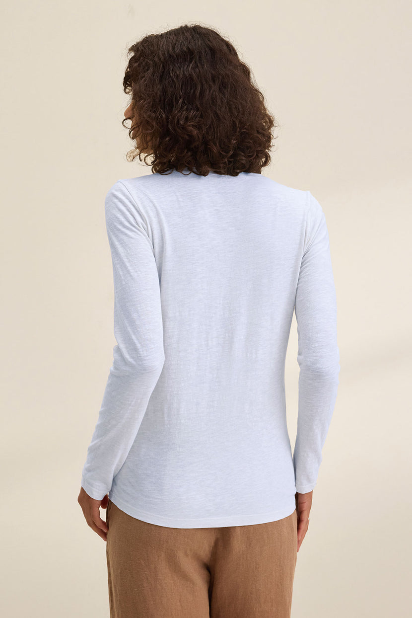 A person with curly hair wears the Velvet by Graham & Spencer BLAIRE TEE, a long-sleeved white v-neck shirt, paired with brown pants and photographed from the back against a neutral backdrop—perfect for effortless everyday style.