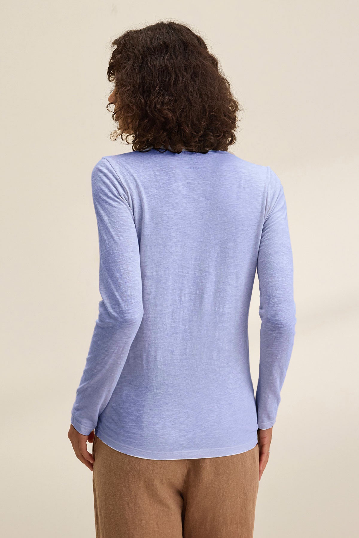 Person with curly hair wears the BLAIRE TEE by Velvet by Graham & Spencer, a light blue cotton slub long sleeve v-neck, paired with brown pants. Shown from the back against a plain background.