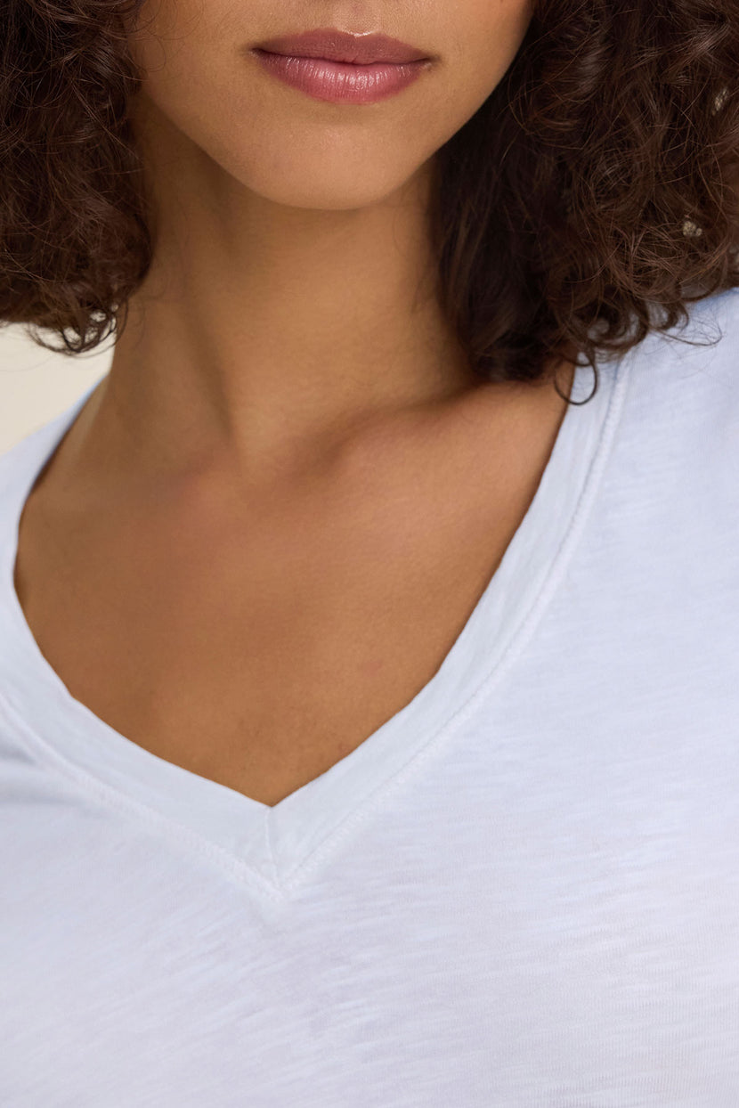 A close-up of a woman in the BLAIRE TEE by Velvet by Graham & Spencer—a white textured cotton slub V-neck shirt—showing her neck, shoulders, and part of her face with curly hair; ideal for effortless everyday style.