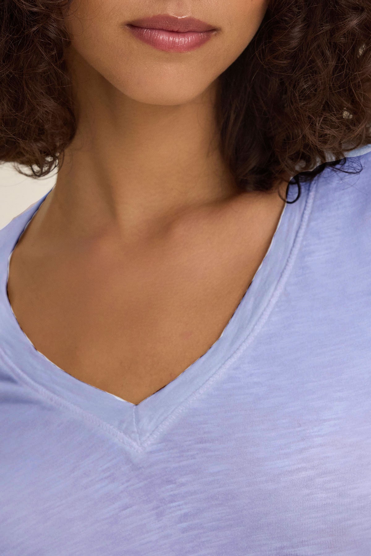 A close-up of a woman wearing the BLAIRE TEE by Velvet by Graham & Spencer in light blue cotton slub with long sleeves and a V-neckline, highlighting her neck, chin, and part of her curly hair.