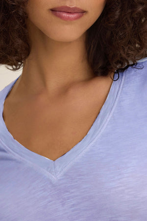 A close-up of a woman wearing the BLAIRE TEE by Velvet by Graham & Spencer in light blue cotton slub with long sleeves and a V-neckline, highlighting her neck, chin, and part of her curly hair.